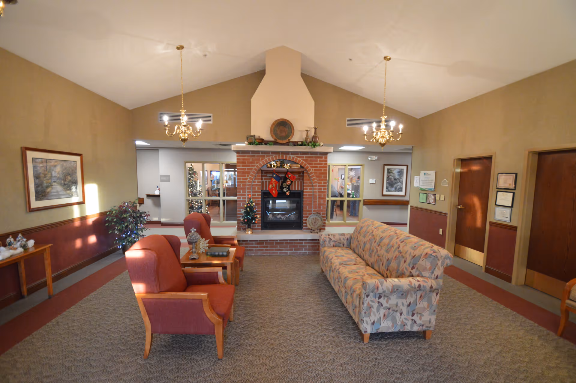 A cozy lounge with upholstered chairs, a patterned sofa, and a central brick fireplace under chandeliers in a senior living facility.