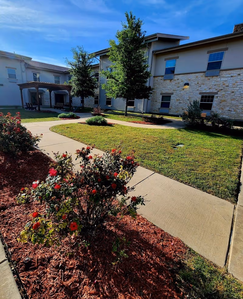 Landscaped courtyard with a paved walkway, flowerbeds and a two-story building under a blue sky.