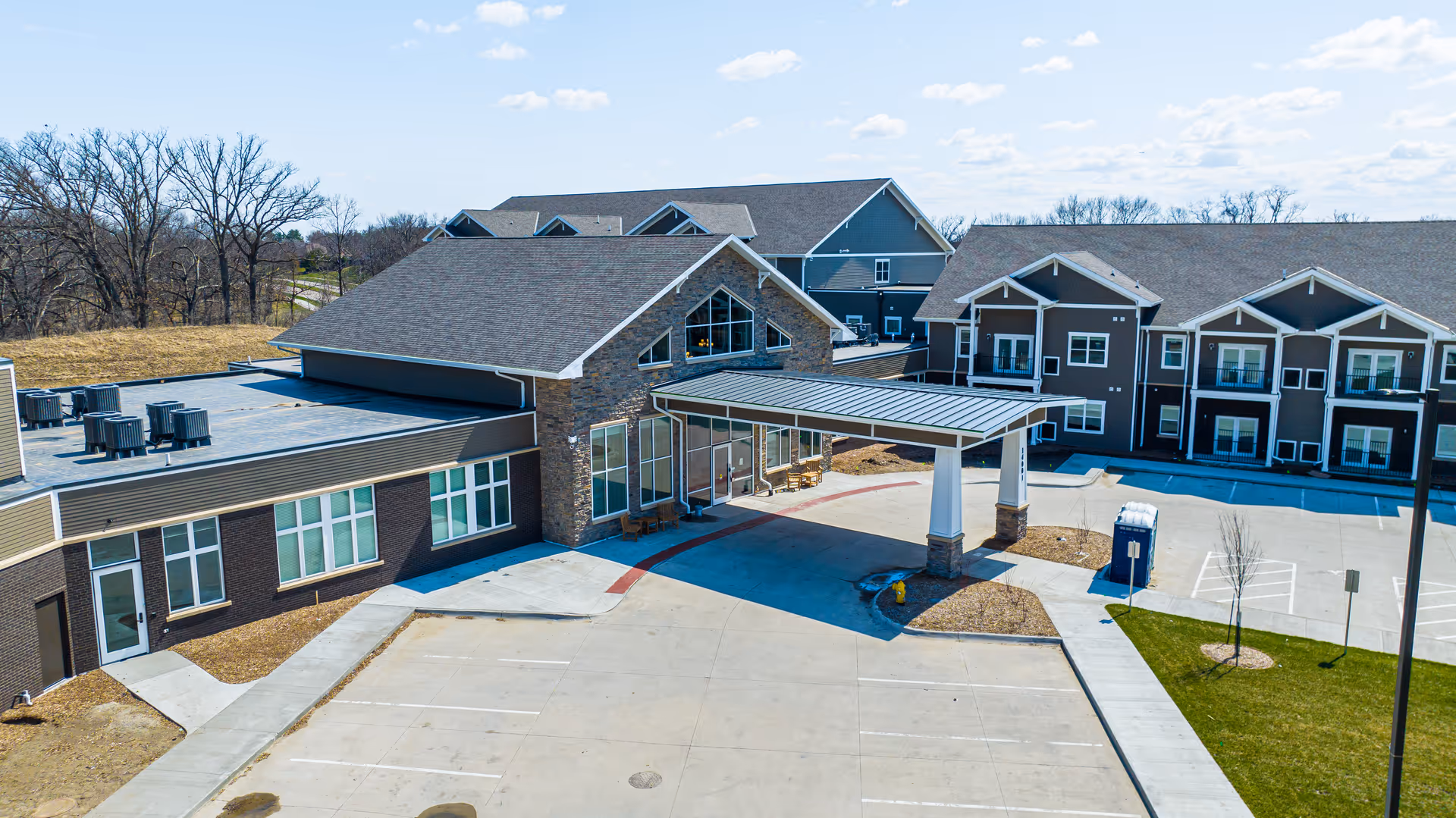 Exterior view of Edencrest at Timberline senior living facility showing multiple connected buildings with pitched roofs, large windows, a covered entrance with stone pillars, and a surrounding parking area with some landscaping and trees in the background under a partly cloudy sky.