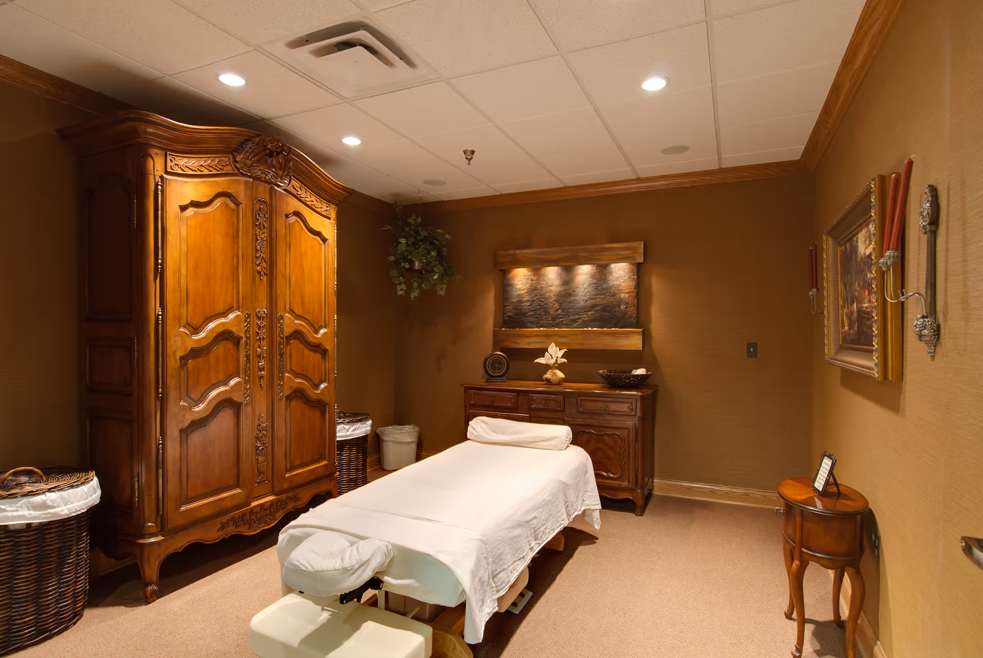 A warmly lit room with a massage table covered in white sheets at the center. The room features a large ornate wooden armoire, a wooden dresser with decorative items, a small round side table, wall sconces with red candles, a framed painting, and a hanging plant in the corner. The walls are brown and the ceiling has recessed lighting.