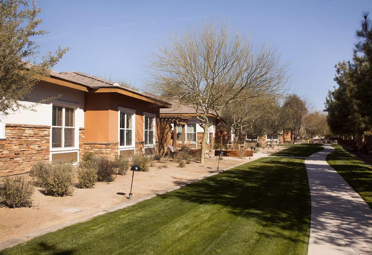 A sunny outdoor pathway lined with green grass and desert landscaping, including bushes and leafless trees, runs alongside a row of single-story buildings with stone and stucco exteriors under a clear blue sky.