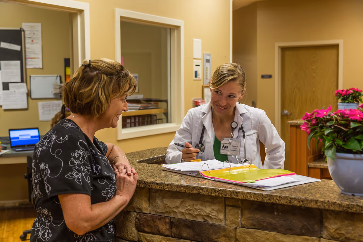 A healthcare professional wearing a white coat and stethoscope is sitting behind a reception desk, smiling and talking to a woman standing on the other side of the desk. The desk has a stone facade and a granite countertop with open binders and paperwork. There are pink flowers in a pot on the desk, and a door and bulletin board are visible in the background.