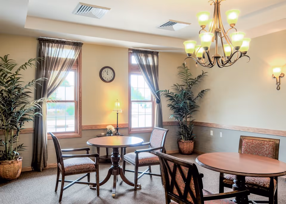 A well-lit room with two round wooden tables, each surrounded by four upholstered chairs. The room has two large windows with sheer curtains, a wall clock between the windows, two potted plants in woven baskets, a table lamp on a small side table, and a chandelier with multiple lights hanging from the ceiling. The walls are painted beige with a wooden chair rail and carpeted floor.