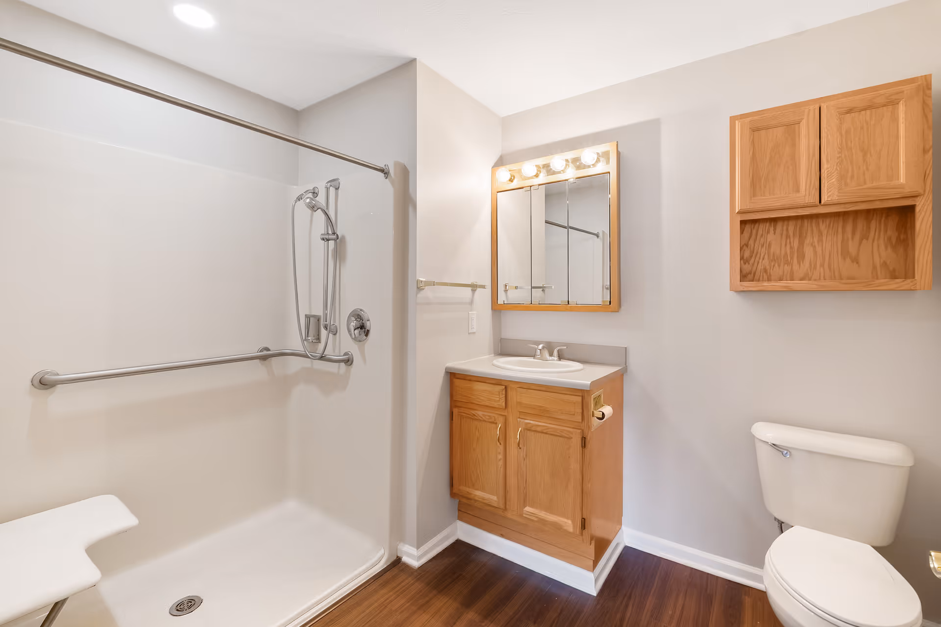 A clean bathroom featuring a walk-in shower with a grab bar and a handheld showerhead, a wooden vanity with a sink and mirror above it, a toilet, and a wooden wall cabinet. The floor is dark wood and the walls are painted light gray.