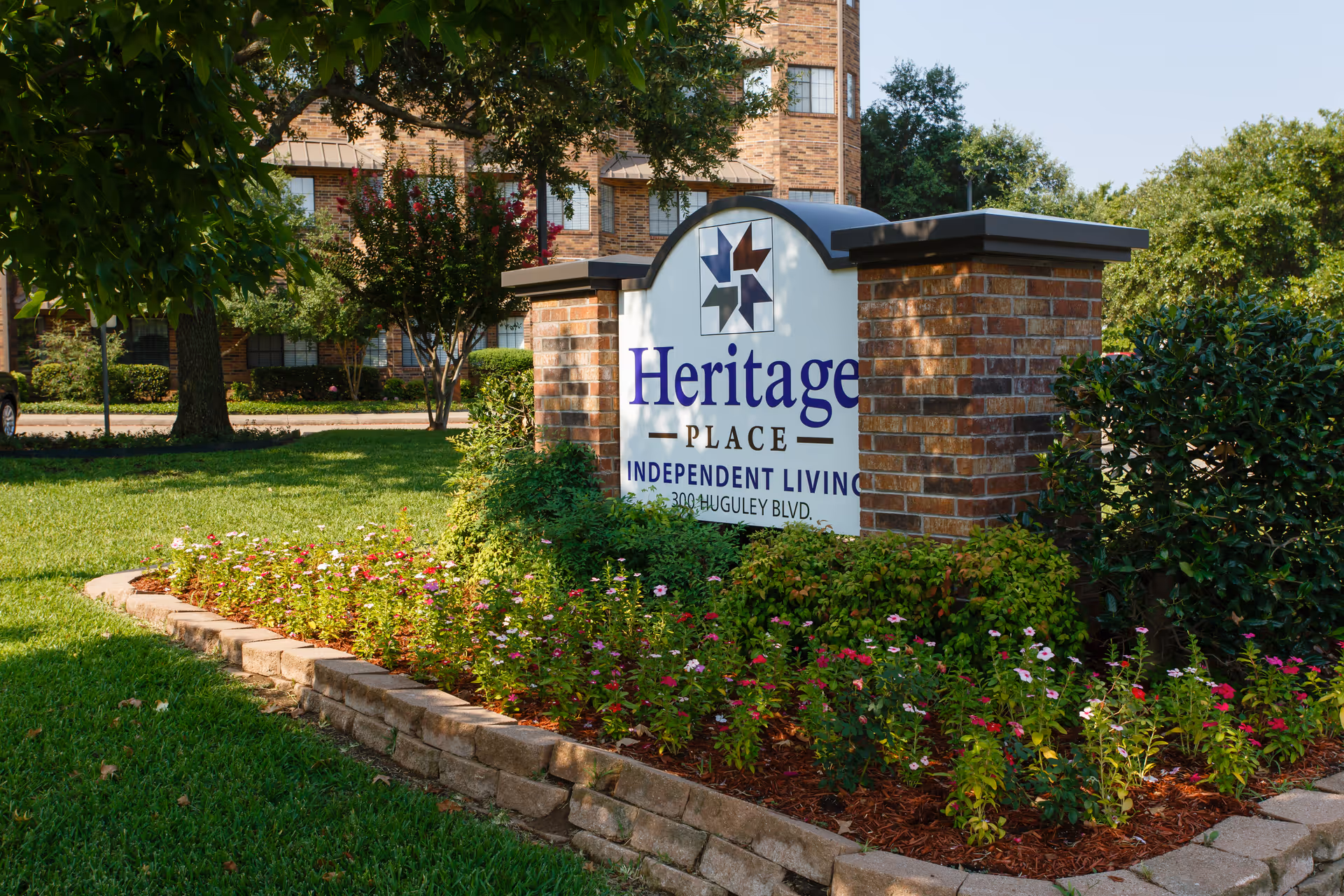 A brick entrance sign reading 'Heritage Place Independent Living' surrounded by flowers and trees in front of a multi-story brick building.