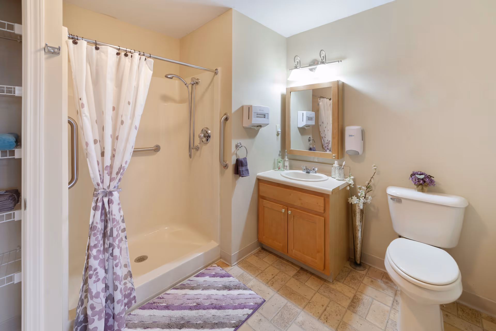 A clean and well-lit bathroom featuring a walk-in shower with a floral-patterned curtain, a wooden vanity with a sink and mirror above it, a toilet with a small flower arrangement on top, and a purple striped bath mat on the tiled floor.