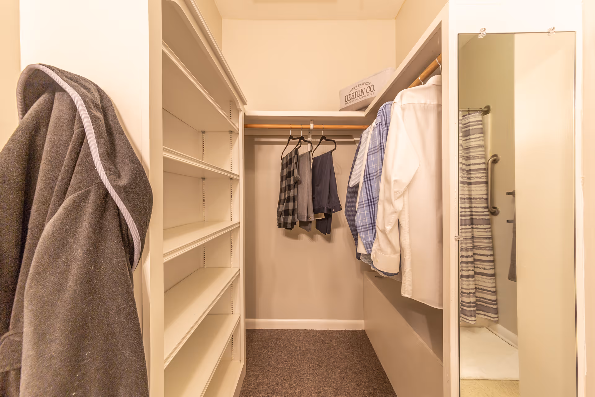 Walk-in closet with shelves on the left, hanging clothes on a rod in the center and right, a full-length mirror on the right wall, and a gray robe hanging on the left side. A glimpse of a bathroom with a striped shower curtain is visible through the mirror reflection.