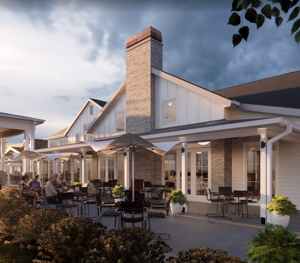 Outdoor patio area of a senior living facility with several tables and chairs under large umbrellas. People are seated and engaged in conversation. The building features a stone chimney, white siding, and large windows with a covered porch. There are potted plants and bushes surrounding the patio.