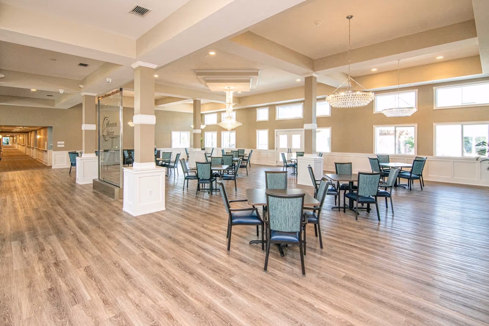 Spacious dining area with multiple tables and chairs arranged on wood flooring. The room features large windows allowing natural light, beige walls with white wainscoting, and elegant chandeliers hanging from a coffered ceiling. A glass panel with the Hampton Manor logo is visible near the entrance.