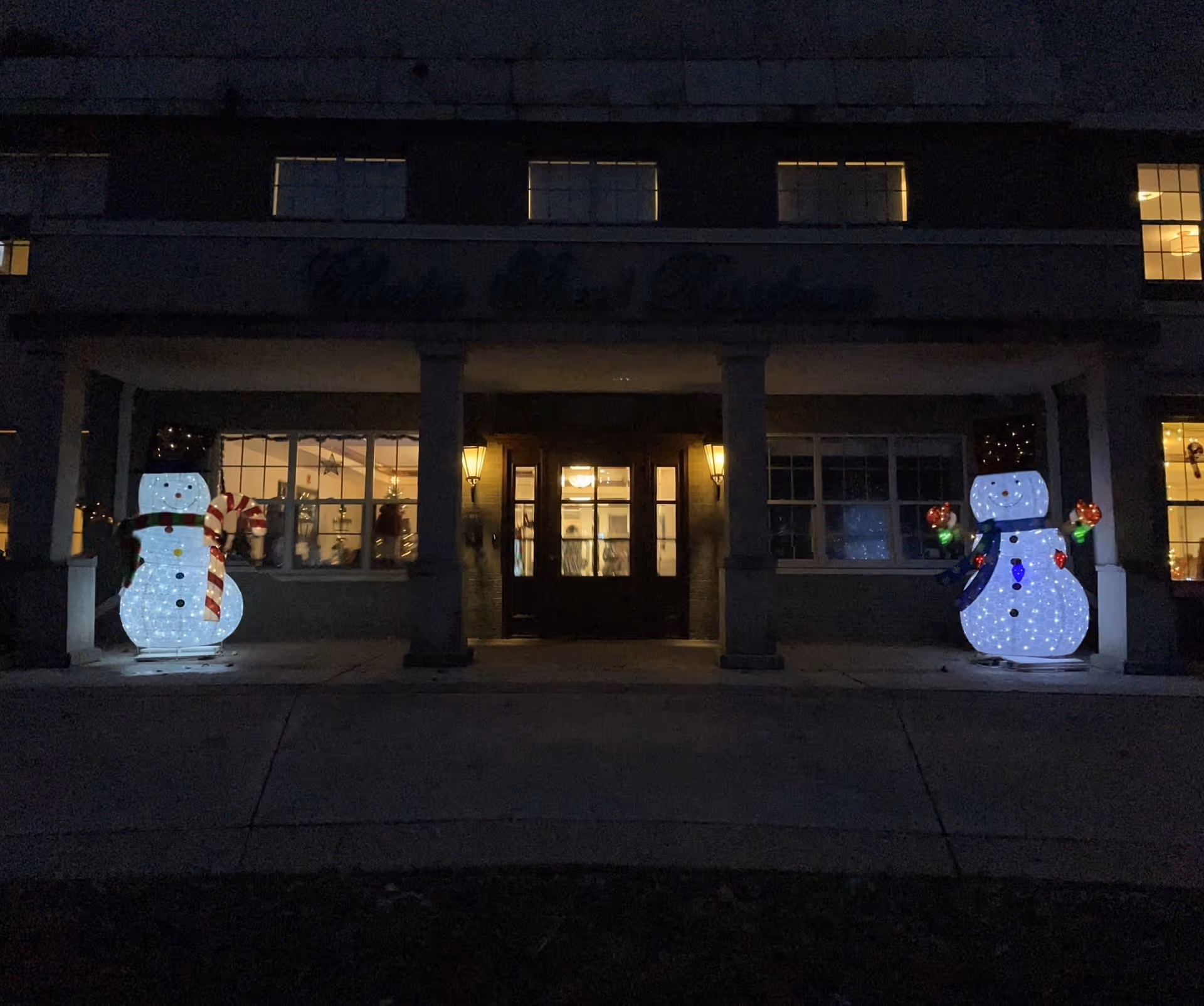 Night view of the entrance to Chester Street Residence decorated with two illuminated snowman figures on either side of the doorway. The building has lit windows and warm exterior lights near the door.