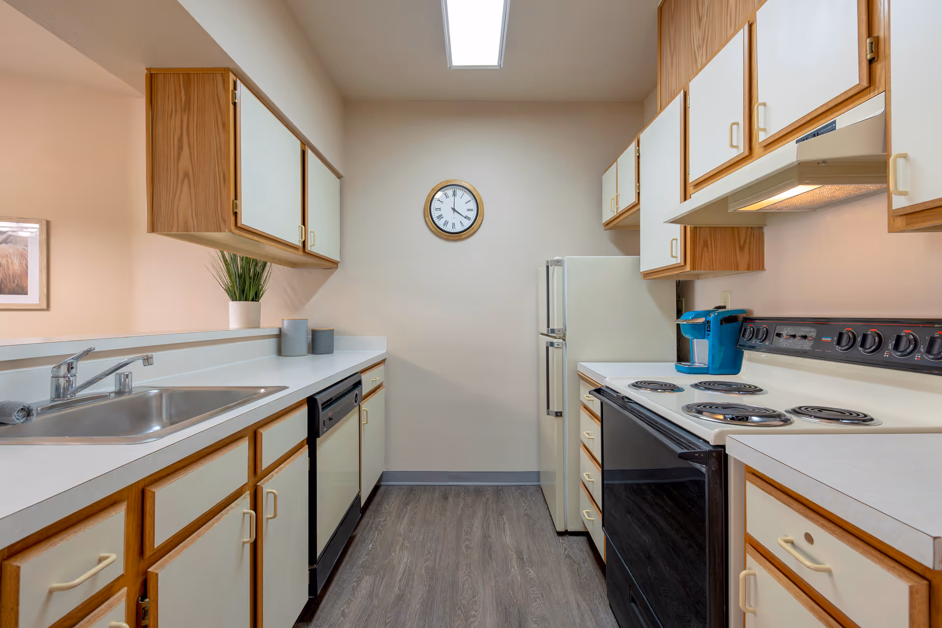 A clean, well-lit kitchen with wooden cabinets and white countertops. The kitchen features a double sink, dishwasher, electric stove with four burners, a refrigerator, and a blue coffee maker. A round wall clock is mounted on the wall at the end of the kitchen.