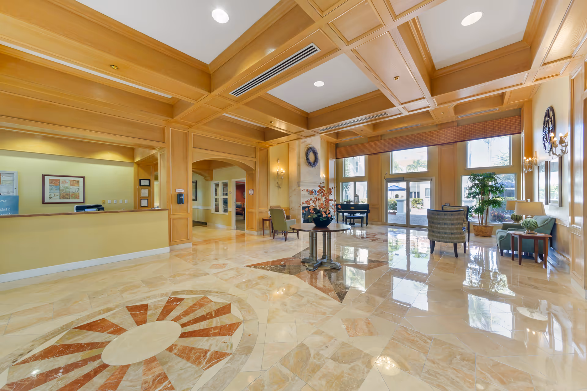 Bright, elegant lobby of a senior living facility with marble floors, a coffered wood ceiling, reception desk, seating areas, and large windows.