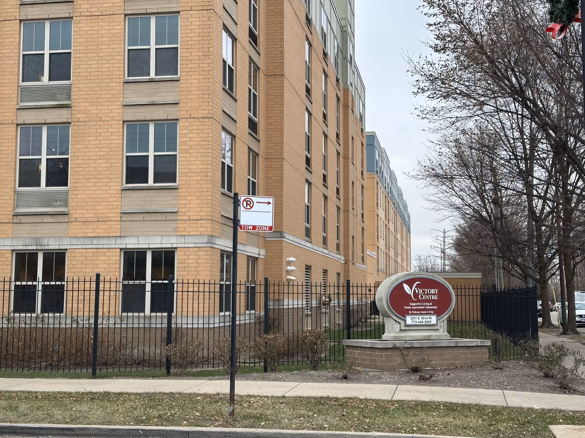 Multi-story brick senior living building with a fenced front yard and a 'Victory Centre' sign by the sidewalk.