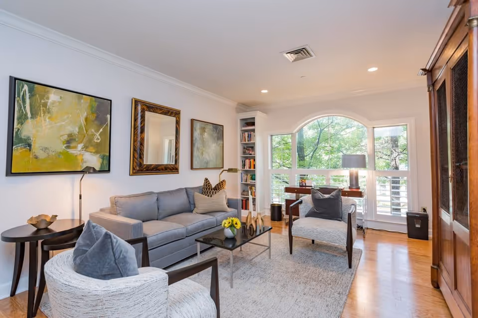 A bright and cozy living room with a large gray sofa, two armchairs with cushions, a glass coffee table with a small vase of yellow flowers, and a side table with a lamp. The room features three framed artworks on the wall, a large arched window with a view of green trees, a wooden cabinet, and a bookshelf filled with books.
