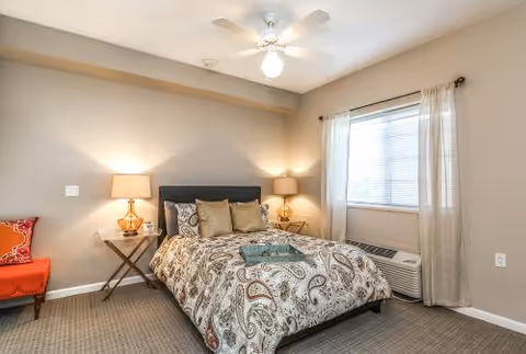 Bright bedroom featuring a patterned bed with pillows, two bedside tables with lamps, a window with curtains, and a ceiling fan.