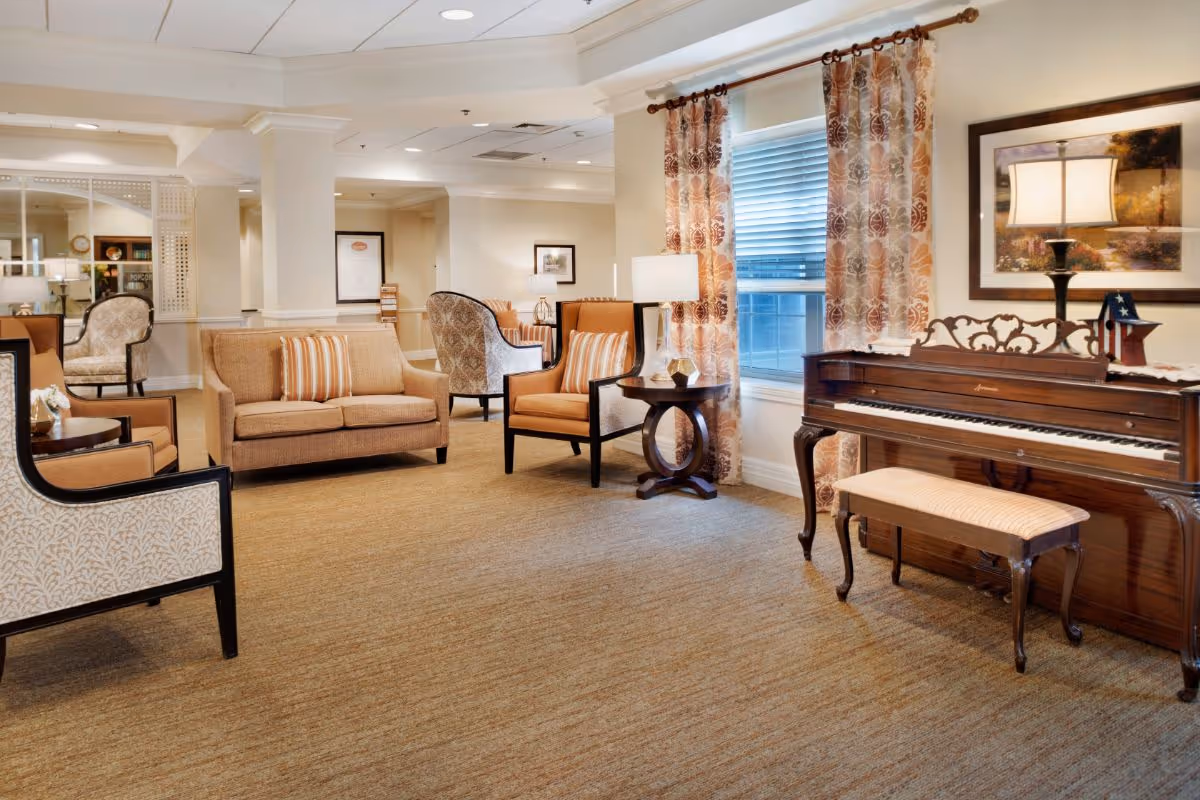 A cozy senior living facility common area with beige carpet, multiple upholstered chairs and sofas with striped cushions, a wooden piano with a matching bench, a side table with a lamp, and a window with patterned curtains. The walls are decorated with framed artwork and the space is well-lit with ceiling lights.