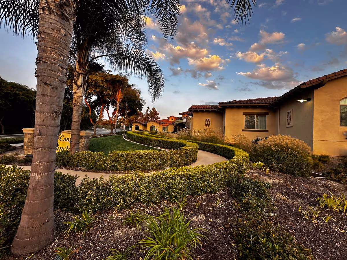 Curved paved walkway and manicured hedges leading past palm trees to single-story stucco buildings under a partly cloudy sky.