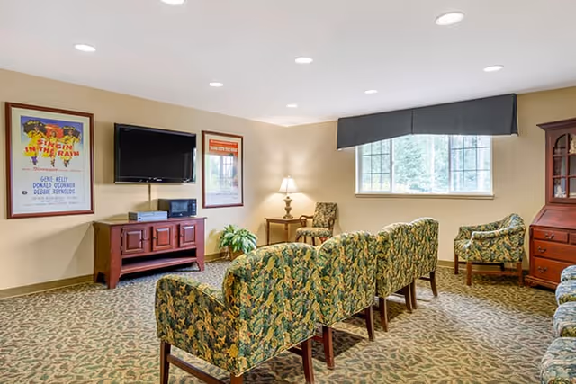 A cozy living room area with patterned upholstered chairs arranged in rows facing a wall-mounted flat screen TV. The room has beige walls, a patterned carpet, a window with a dark valance, a wooden cabinet, a small side table with a lamp, and framed posters on the wall.