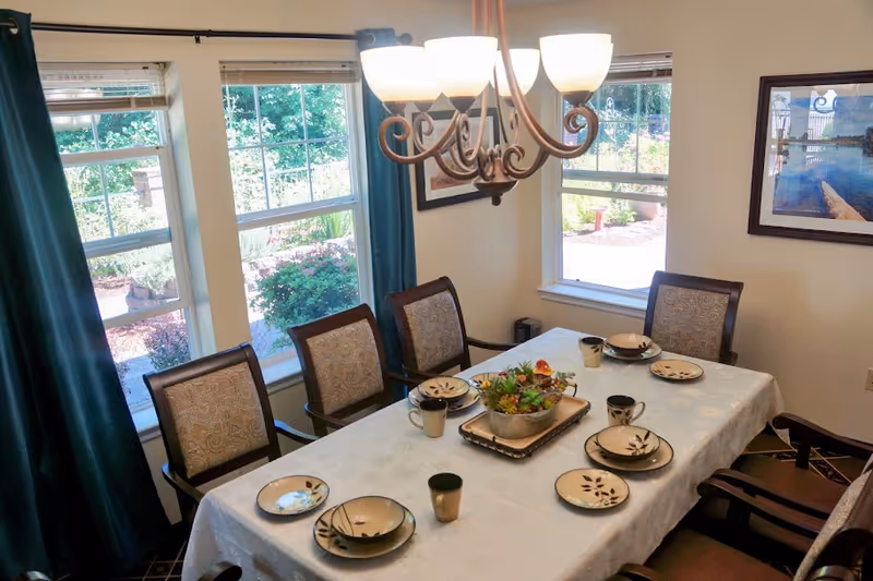 A dining room with a rectangular table covered with a white tablecloth. The table is set with plates, bowls, mugs, and a centerpiece with green plants. There are six upholstered chairs around the table. Three large windows with blue curtains let in natural light, and a chandelier with five lights hangs above the table. A framed picture is on the wall to the right.