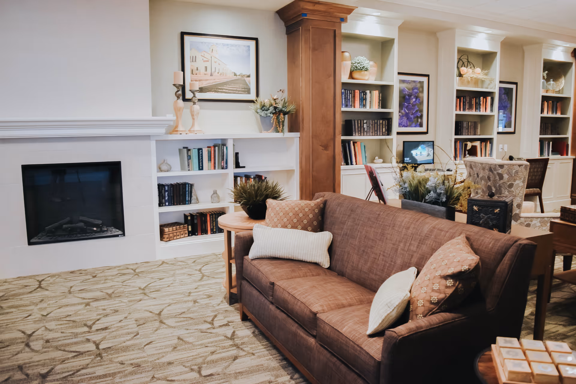 A cozy senior living facility common area featuring a brown sofa with patterned and plain cushions, a fireplace with a white mantel, built-in bookshelves filled with books and decorative items, framed artwork on the walls, and a carpeted floor with a subtle pattern.