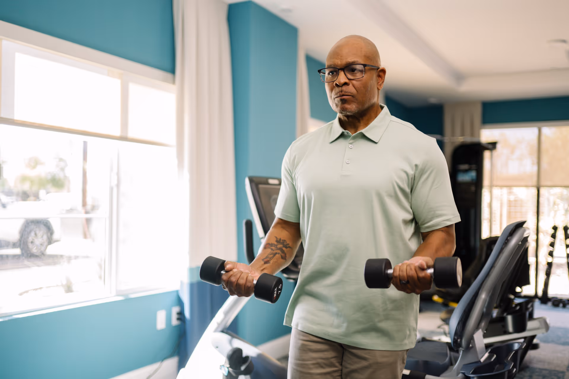 An older man wearing glasses and a light green polo shirt is lifting dumbbells in a well-lit exercise room with large windows and various fitness equipment.