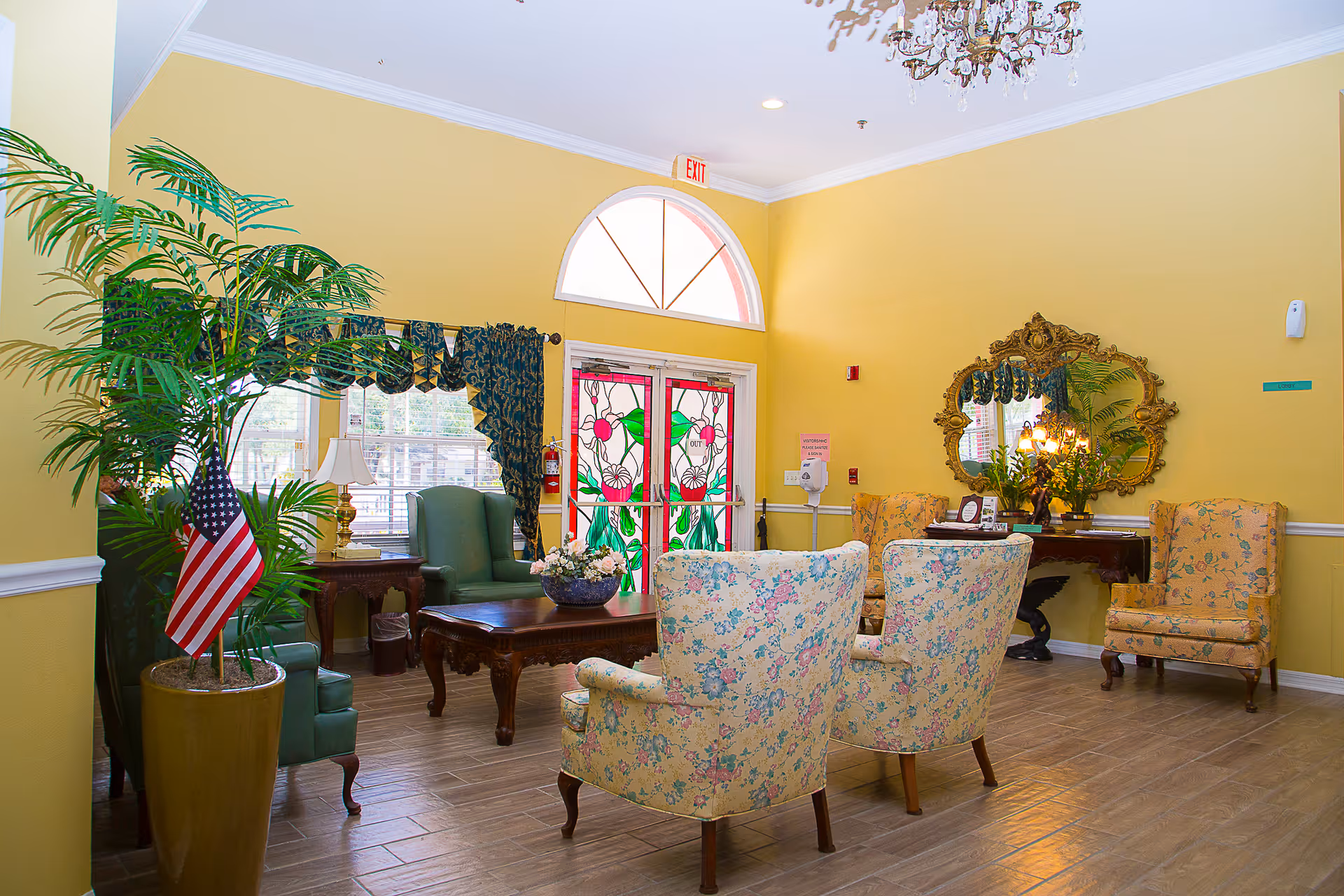 A bright and welcoming assisted living facility common area with yellow walls, floral upholstered chairs, a wooden coffee table with a flower arrangement, green armchairs, a large potted plant with an American flag, a decorative mirror above a console table with flowers, and stained glass double doors with an exit sign above.