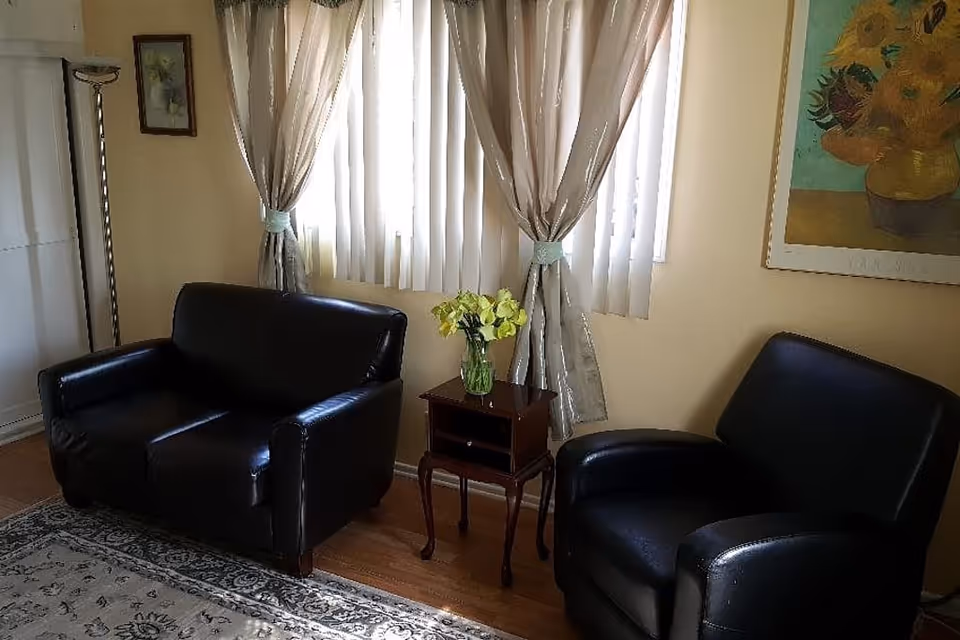 Small living room with two black leather chairs, a wooden side table holding a vase of flowers, and curtained windows.