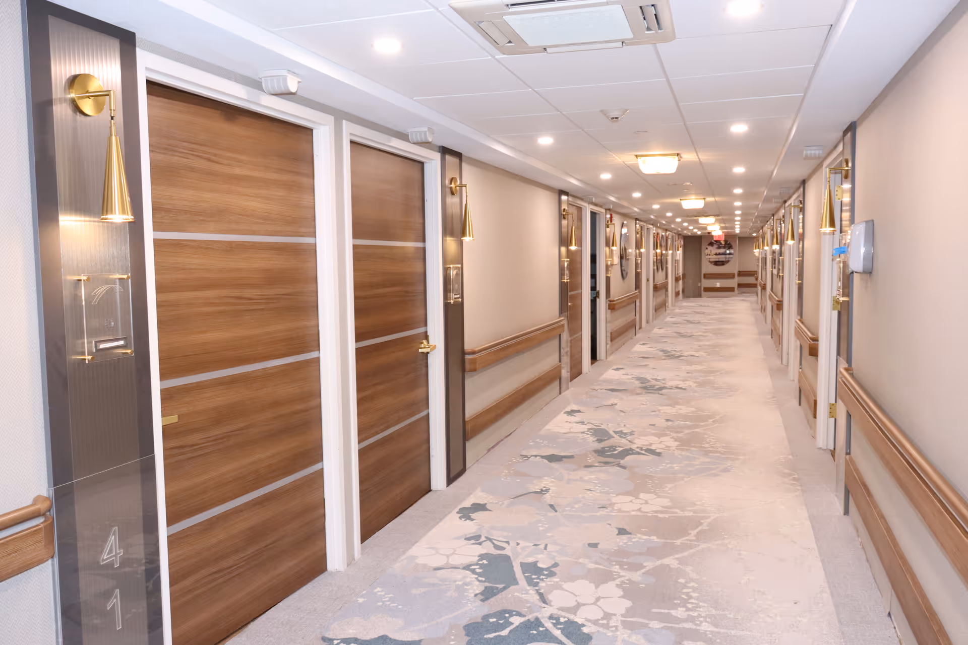 Long well-lit interior hallway with wooden room doors, handrails, and a patterned carpet in a senior living facility.