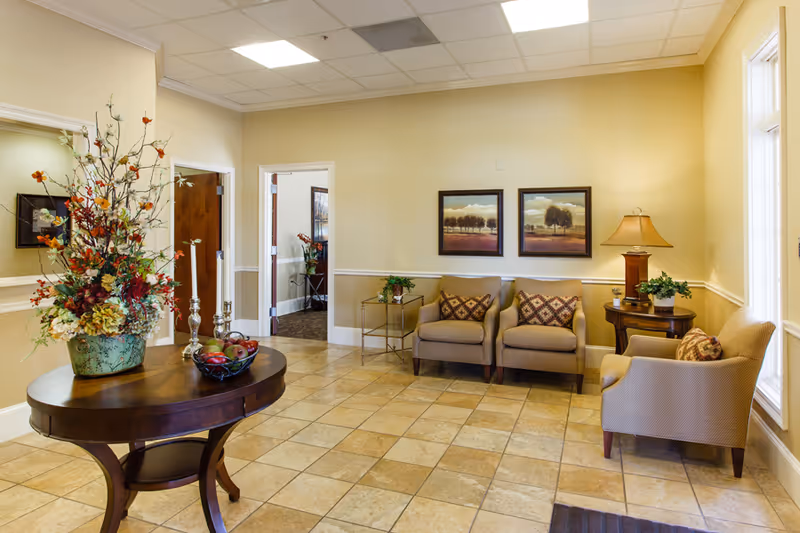 A cozy sitting area in an assisted living facility with beige walls and tiled floor. The room features a round wooden table with a large floral arrangement and decorative items, two beige armchairs with patterned cushions, a small side table with a lamp and plant, and two framed landscape paintings on the wall. There is a window letting in natural light and an open doorway leading to another room.