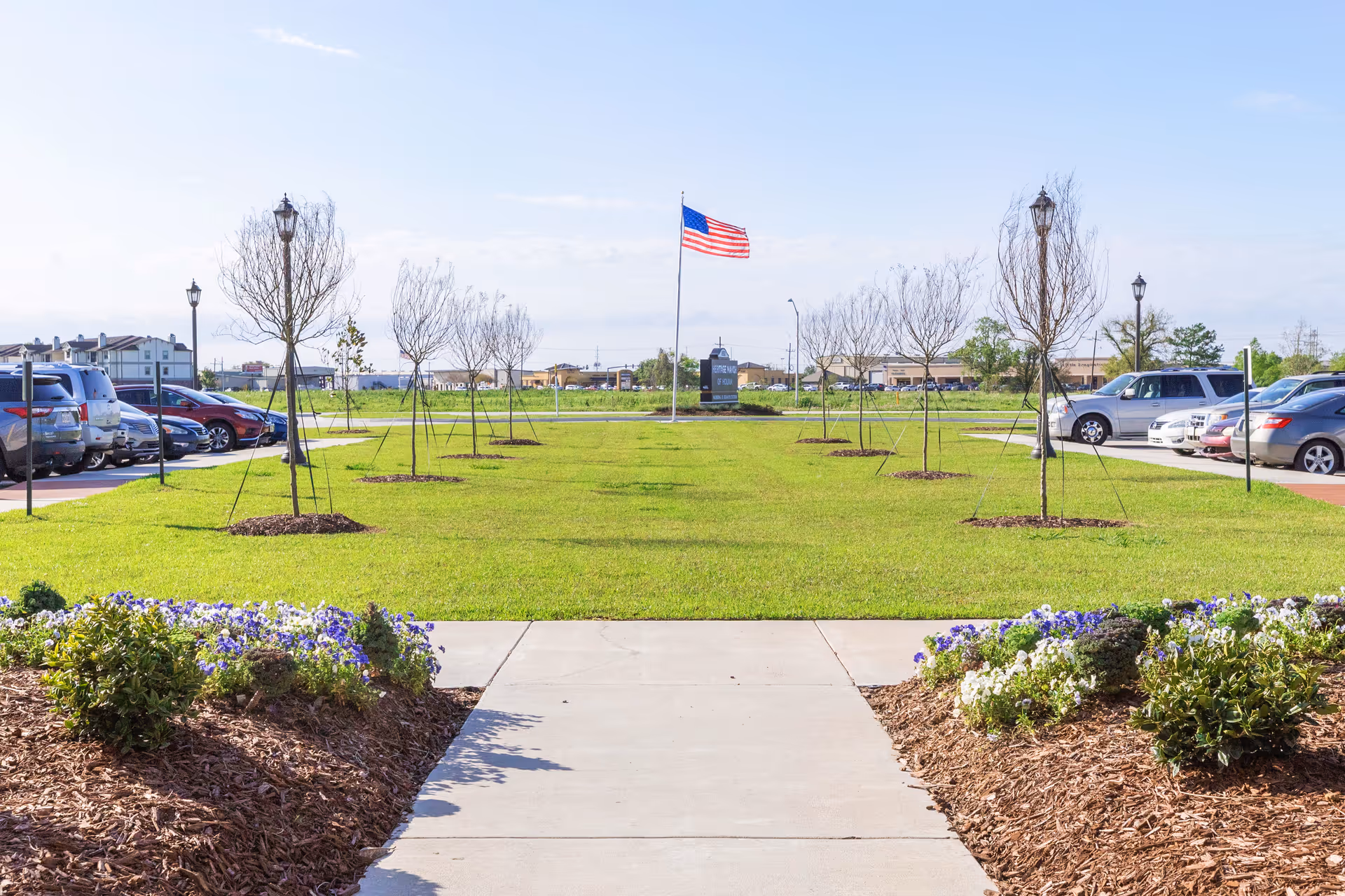 A landscaped outdoor area with a concrete walkway leading to a grassy lawn lined with young trees and flower beds on either side. An American flag is flying on a flagpole in the center background, with parked cars visible on both sides of the lawn under a clear blue sky.