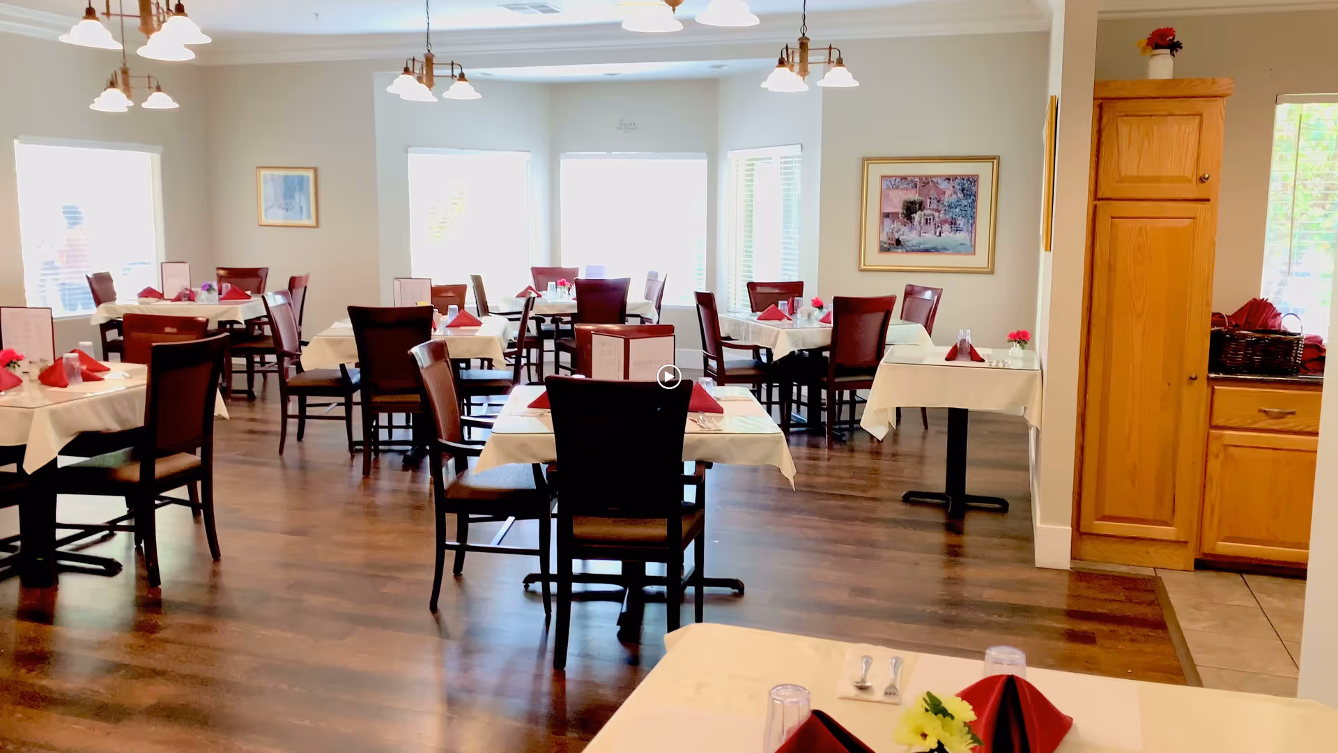 A bright dining room with multiple tables covered in white tablecloths and set with red napkins, menus, and small flower arrangements. The room has wooden floors, large windows letting in natural light, and light fixtures hanging from the ceiling. There is a wooden cabinet on the right side near a window.