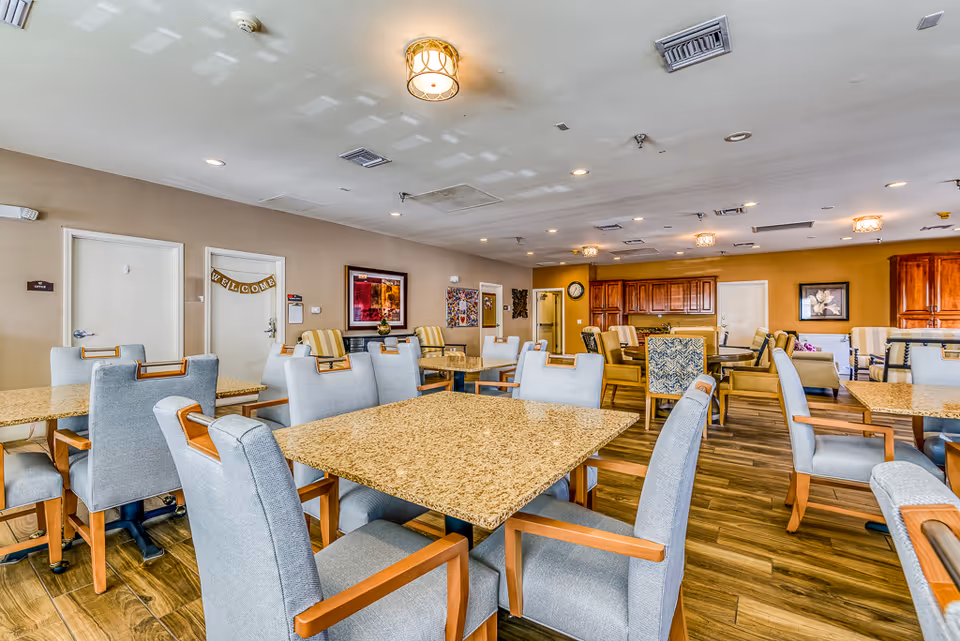 Spacious communal dining room with multiple tables and upholstered chairs in a senior living facility.