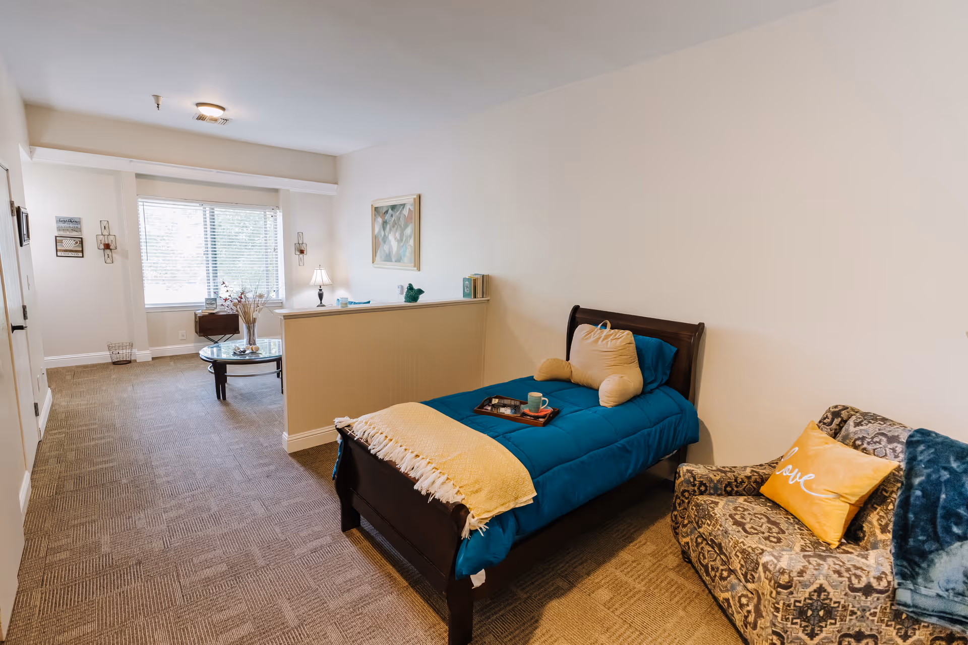 A cozy bedroom in an assisted living facility featuring a single bed with blue bedding and a yellow throw blanket, a decorative pillow shaped like a bone, and a tray with a cup and book on the bed. Next to the bed is a patterned armchair with a yellow pillow and a blue throw blanket. The room has beige carpet, a half wall separating the bed area from a sitting area with a round glass coffee table and a window with blinds letting in natural light. A lamp and framed artwork are also visible.