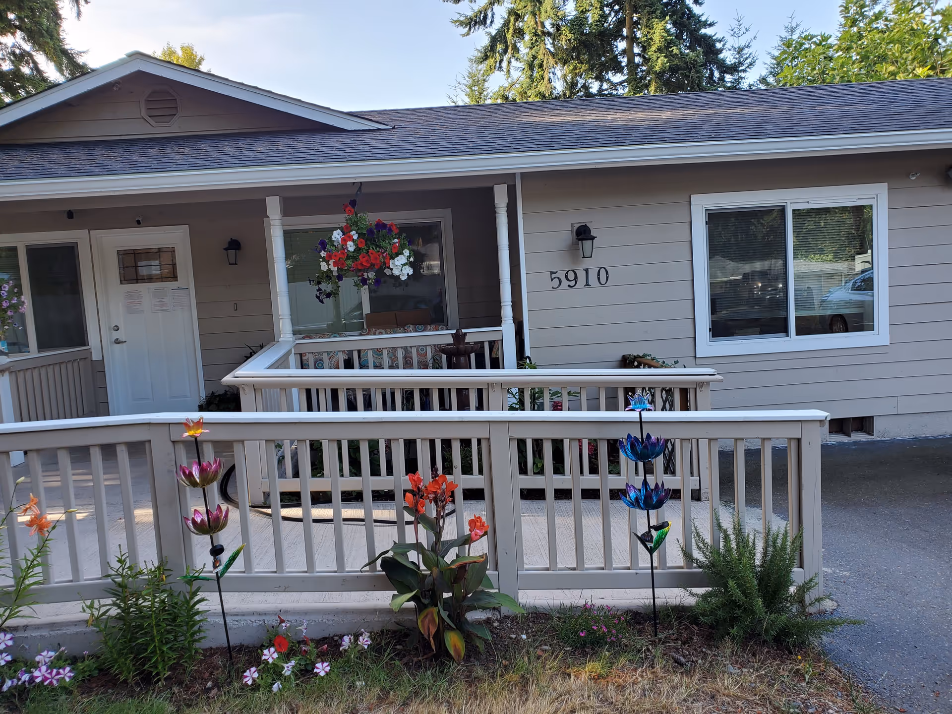 Front exterior view of a single-story building with the address number 5910 displayed on the wall. The building has a small porch with white railings and hanging flower baskets. There are colorful decorative flowers planted in the garden bed in front of the porch. Trees and greenery are visible in the background.