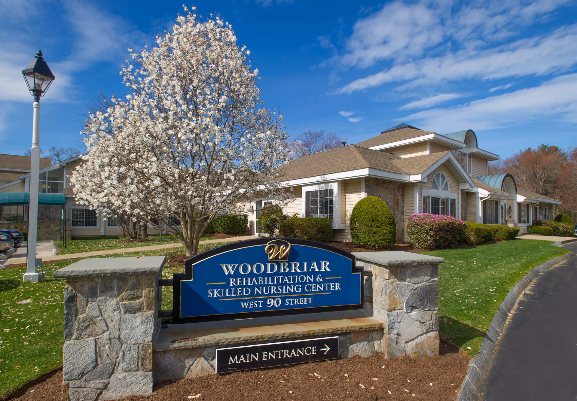 Exterior view of Woodbriar Rehabilitation & Skilled Nursing Center with a stone sign in front, a blooming tree, well-maintained bushes, and a clear blue sky.