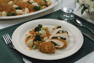 A white plate with sliced breaded chicken stuffed with spinach, served with mixed steamed vegetables including broccoli, cauliflower, and carrots. The plate is on a green placemat with a fork to the left and a knife and spoon to the right. In the background, there is a glass of water, a small flower arrangement, and a serving dish with more breaded chicken.