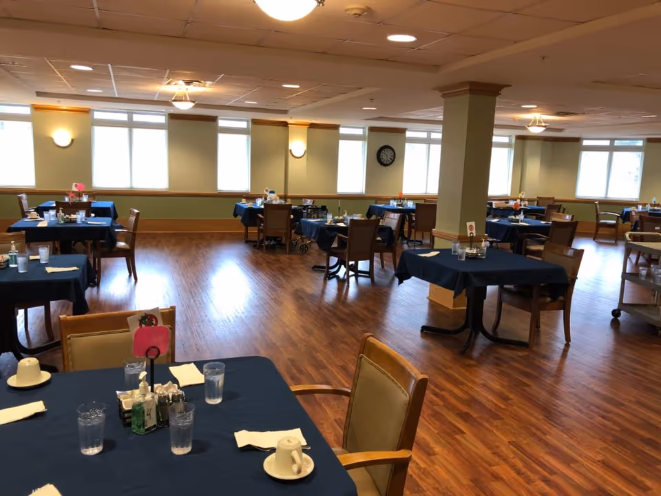 A spacious dining room with multiple tables covered in dark blue tablecloths, each set with glasses, cups, napkins, and condiments. The room has wooden flooring, large windows letting in natural light, and a clock on the wall. Chairs are arranged around the tables, and the overall atmosphere is clean and orderly.