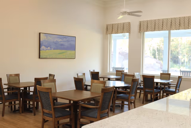 A bright dining room with multiple wooden tables and chairs arranged neatly. Large windows allow natural light to fill the room, and a landscape painting hangs on the wall. The room has a clean, welcoming atmosphere with light-colored walls and a ceiling fan.