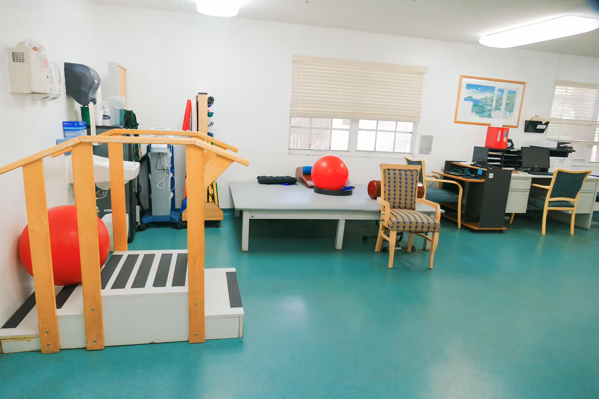 A rehabilitation or physical therapy room with a small wooden staircase with handrails, a large red exercise ball, a padded therapy table, two patterned chairs, and a desk with two computer monitors. The room has teal flooring, white walls, and windows with blinds. There is a framed picture on the wall above the desk.