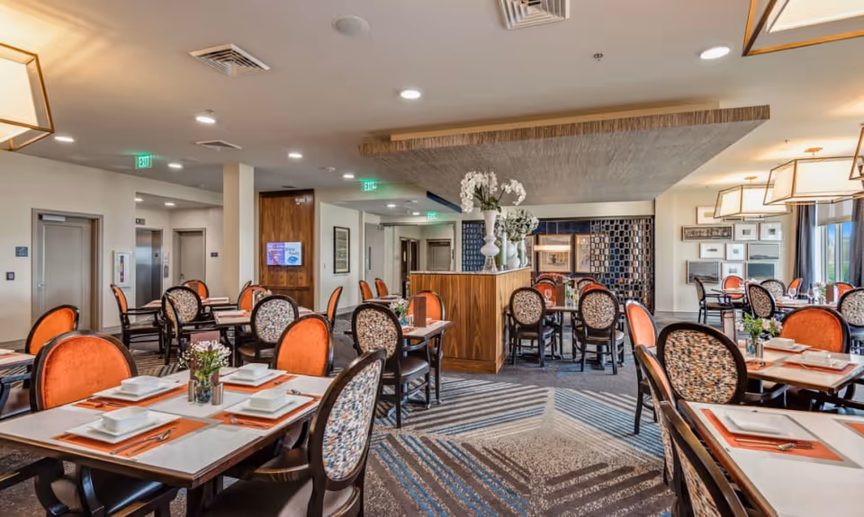 A bright and spacious dining room in a senior living facility with multiple tables set with white plates, bowls, and orange placemats. The chairs have patterned backs and orange cushions. The room features modern lighting fixtures, decorative vases with flowers, and large windows allowing natural light to enter.