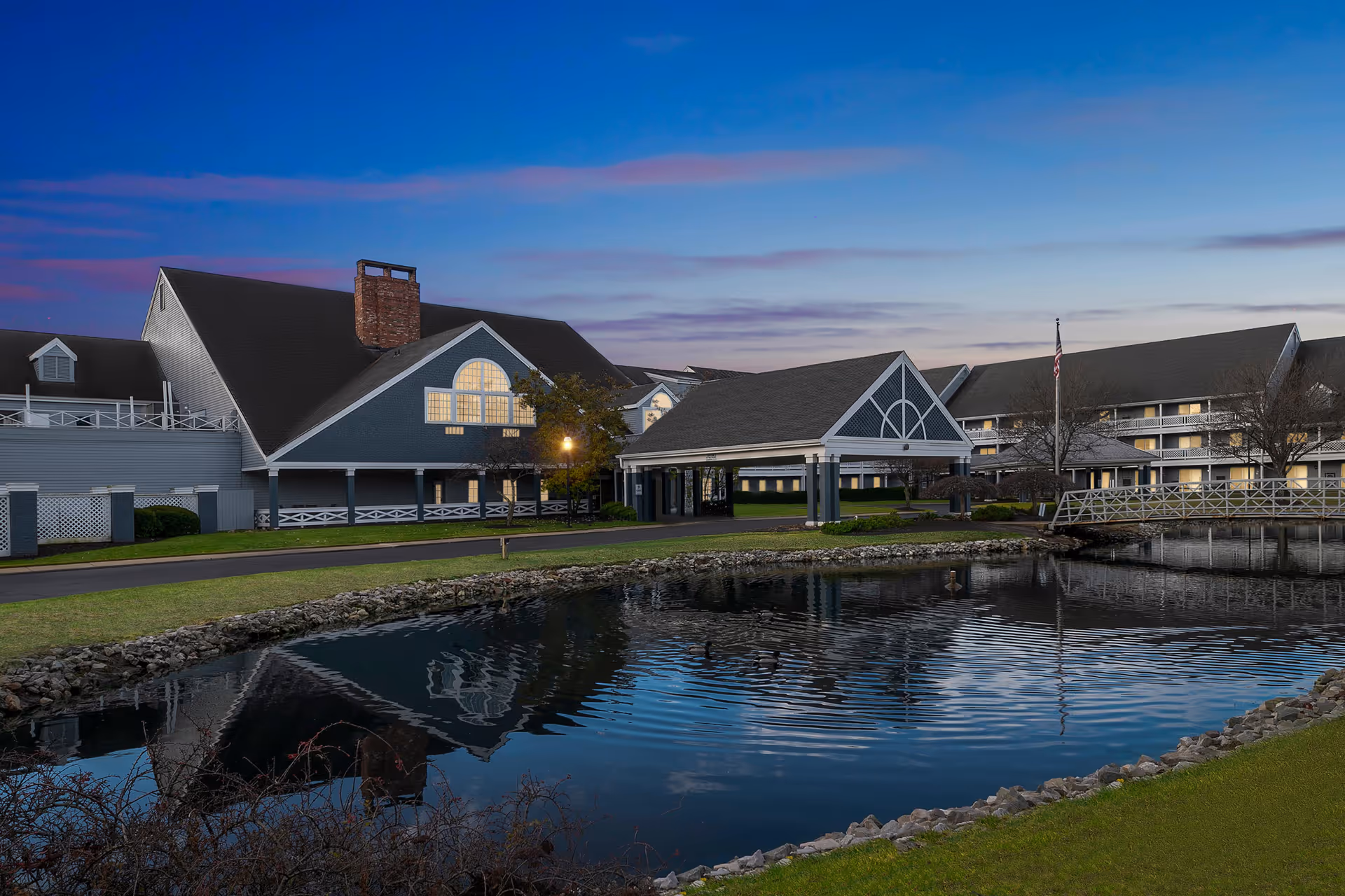 Exterior view of a senior living facility at dusk with a large pond in the foreground reflecting the buildings. The buildings have pitched roofs, large windows, and a covered entrance. There is a small bridge over the pond and an American flag on a flagpole near the entrance.
