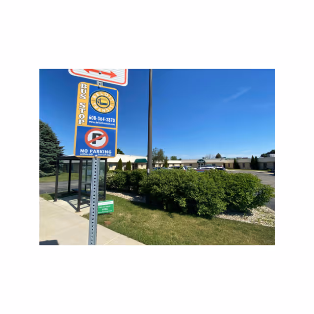 Outdoor view of a bus stop sign near a sidewalk with a shelter, green bushes, and a low building in the background under a clear blue sky.
