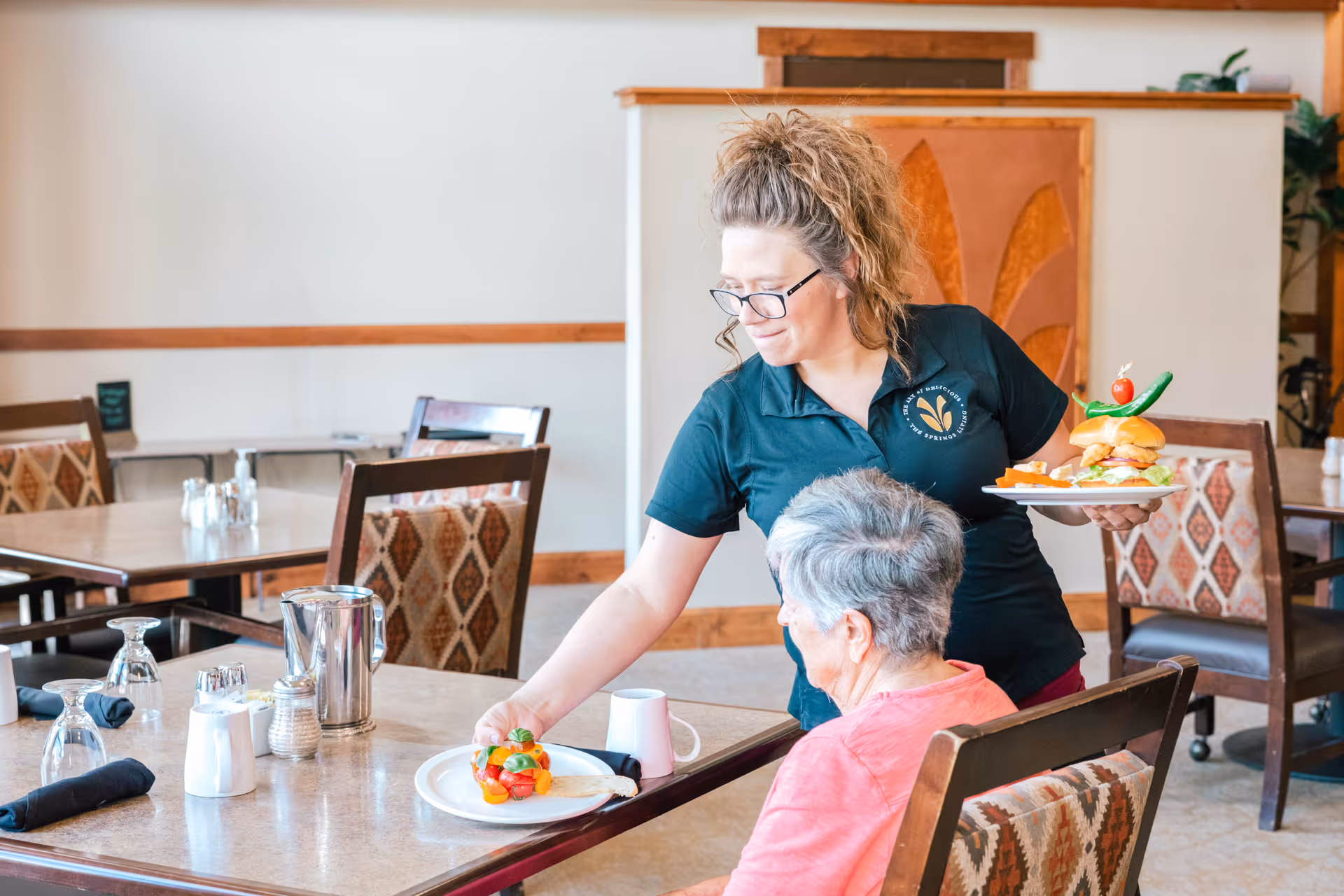 A dining room in The Springs at Butte facility with a staff member serving food to an elderly woman seated at a table. The staff member is holding a plate with a sandwich and placing another plate with a colorful salad on the table. The room has wooden chairs with patterned upholstery and tables set with glasses and napkins.