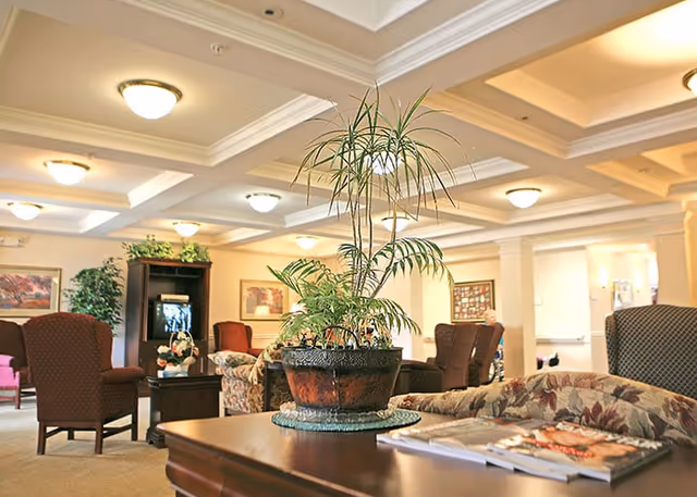 A well-lit senior living facility common area with comfortable upholstered chairs and sofas arranged around a central coffee table. A large potted plant sits on a wooden table in the foreground, with magazines placed nearby. The ceiling features recessed lighting and decorative coffered panels. Artwork and additional plants decorate the walls and corners of the room.