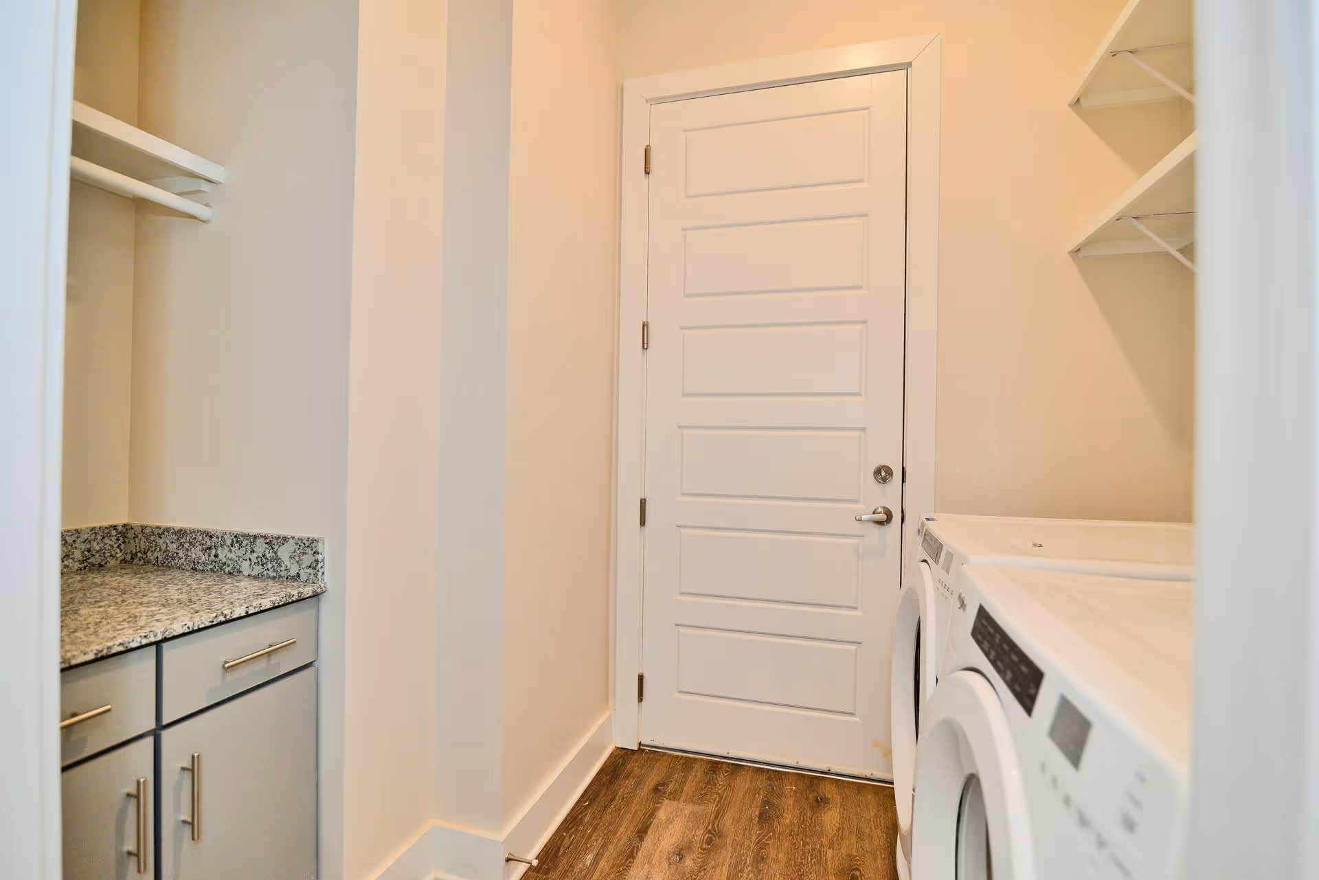 Small laundry room with front-loading washer and dryer, granite countertop, cabinets, shelving, and a closed white door.