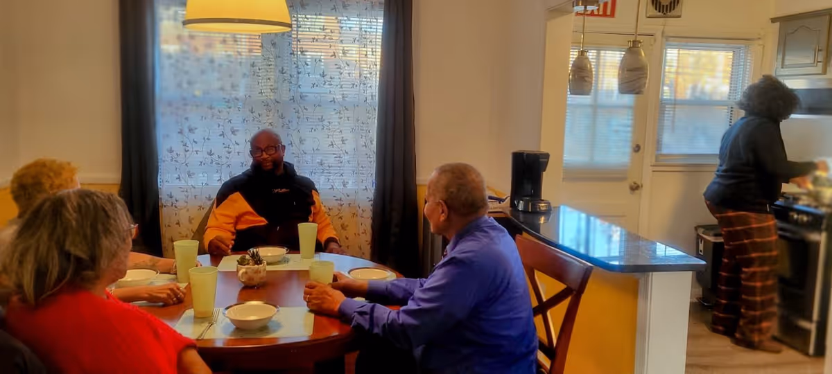 Four people sitting around a dining table with bowls and cups, engaged in conversation. In the background, a person is standing and cooking in a kitchen area with a stove and countertop. The room has a window with sheer curtains and pendant lights above the kitchen counter.