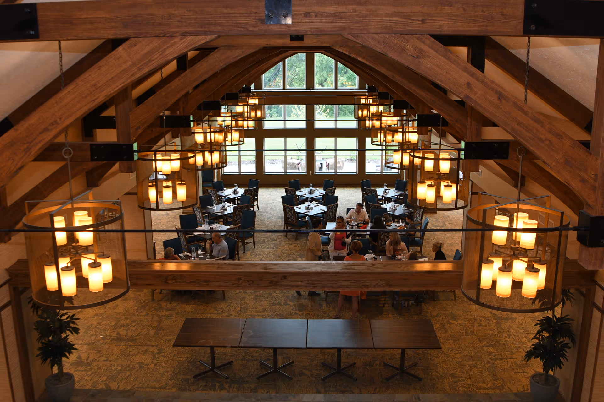 Spacious wood‑beamed dining room with large chandeliers and tables where residents are seated near floor‑to‑ceiling windows.
