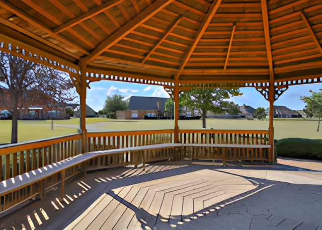 View from inside a wooden gazebo with built-in benches, overlooking a grassy area with trees and houses in the background under a blue sky.
