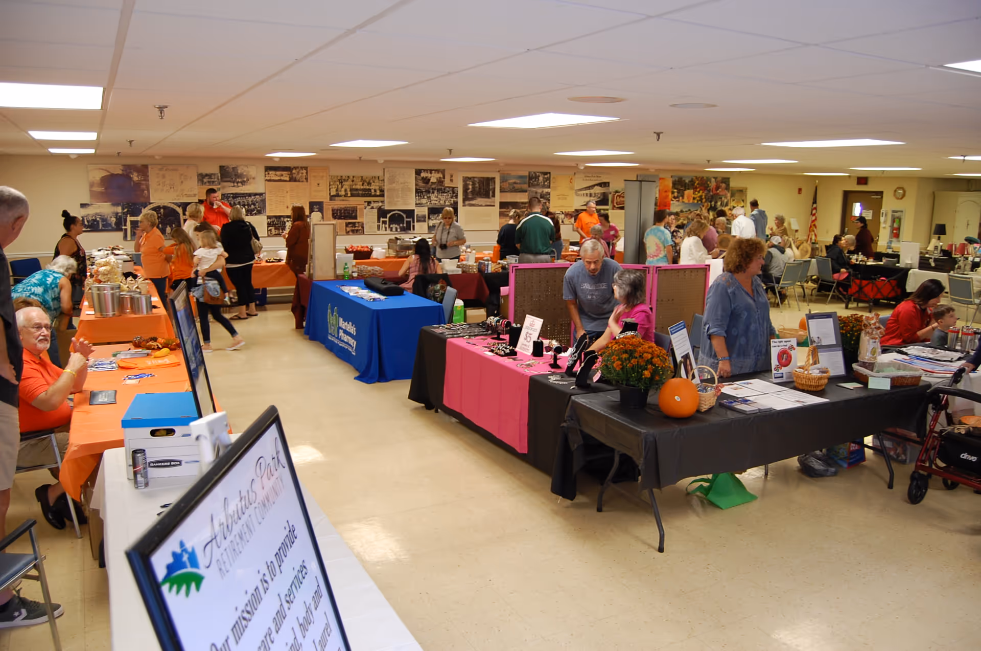 Indoor community event with multiple tables covered in colorful tablecloths displaying various items and information. People are standing and sitting around the tables, engaging in conversations and browsing. The room has a tiled floor, fluorescent ceiling lights, and historical photos on the far wall.