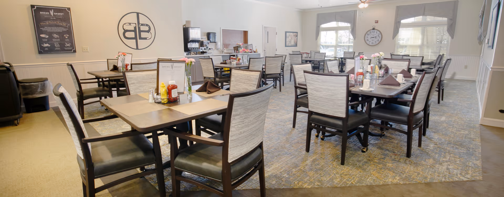 Spacious dining room with multiple tables and chairs set for meals in a senior living facility.